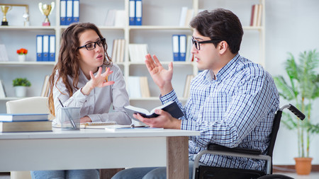 Ein Student im Rollstuhl und eine Studentin besprechen am Tisch mit Büchern eine Aufgabe