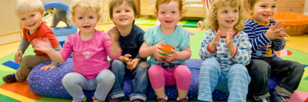 Sechs Kleinkinder sitzen lachend und klatschend auf einem bunten Kissen